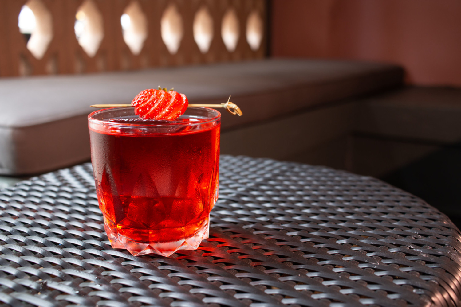 A red drink in a clear glass topped with sliced strawberries. It is photographed on a brown woven table.