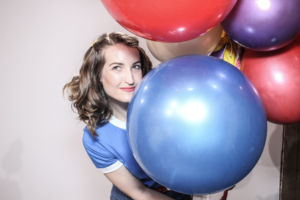 A woman poses with brightly colored balloons. She is wearing a blue shirt while looking away from the camera.