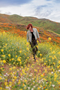 A woman wearing black shirt and pants with a jean jacket walking through a field of yellow flowers.
