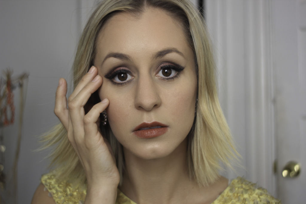 A blonde woman wearing yellow. She is looking at the camera, her right hand touching her face.