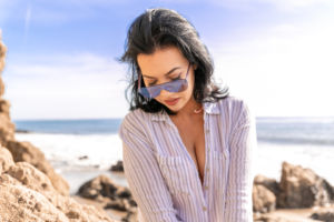 A woman with pair of sunglasses on her nose looking down, she is wearing a striped shirt. Her background is a blurred beach.