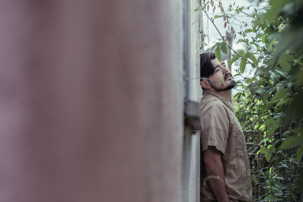 Wearing a cream shirt, a man is leaning on a peach colored wall while looking at a distance. In front of him are tall plants.