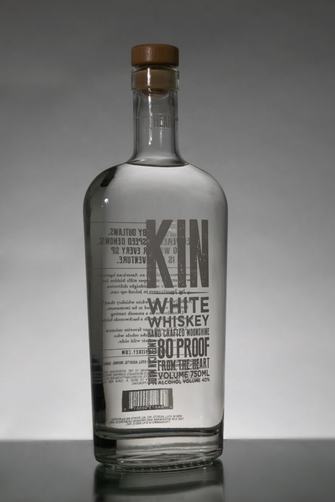 A Kin White Whiskey bottle photographed on a gray surface and white wall.