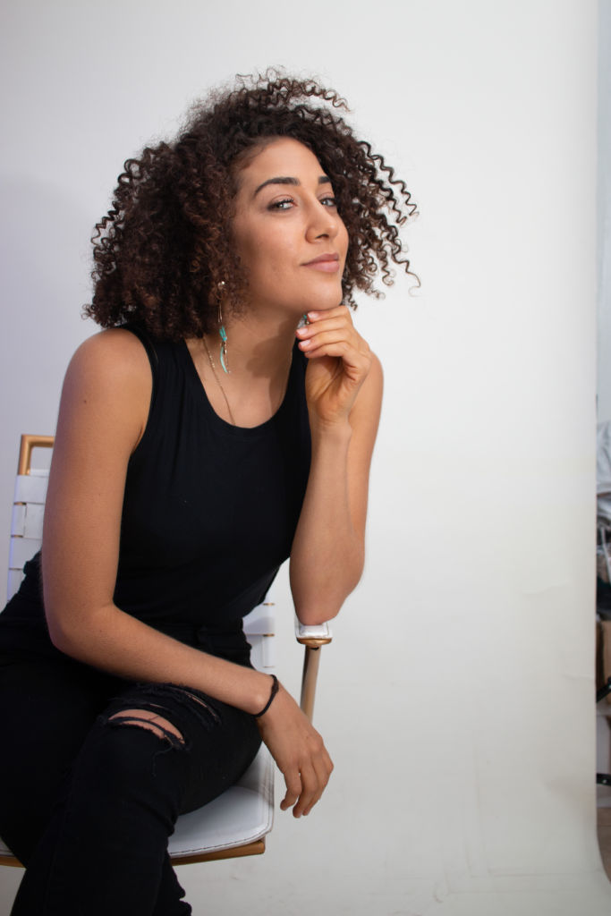 A woman dressed in a black tank top and black ripped pants smiling at the camera while sitting on a white seat.