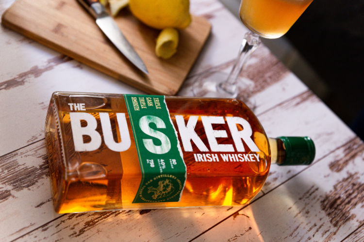 Busker Whiskey photographed with a tall glass, there is a whole lemon, a few slices , a small knife, and a wooden cutting board on a wooden background.