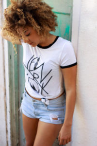 A woman looking downwards, wearing a white shirt with a New York print and jean shorts.
