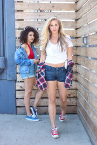 Two girls looking at a photo. One girl is leaning on the wooden wall, wearing a jean jacket and shorts with a red top. The second girl is walking towards the camera, wearing a white top and jean shorts with her plaid shirt hanging on her arms.