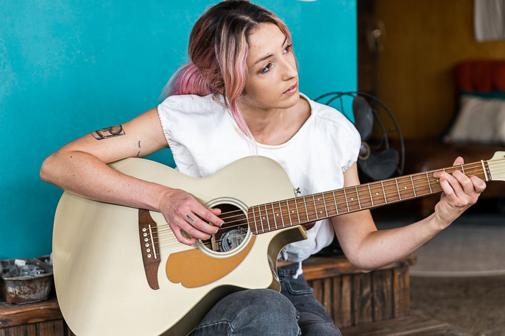 Wearing a white top and jeans, a young woman playing a guitar as she gazes away from the camera.
