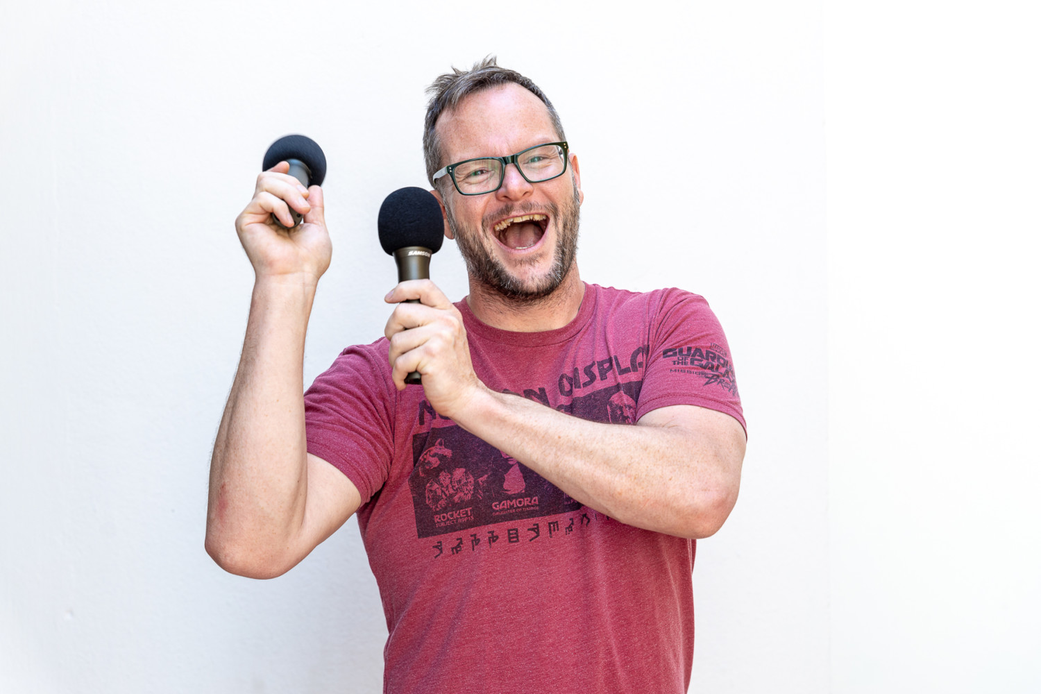 A young man in glasses holds two microphones with a wide smile.