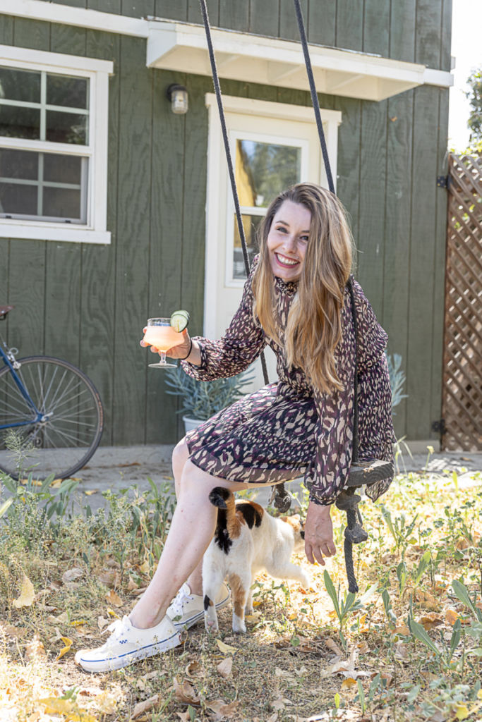 A young woman smiling at the camera as she is perched on a swing with a drink in hand and a cat by her leg.