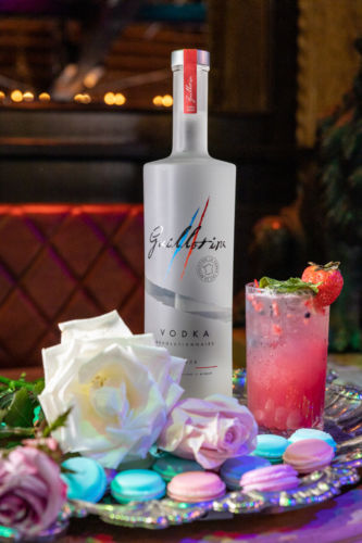 Guillotine vodka photographed with a pink drink, macarons, and flowers on a tray.