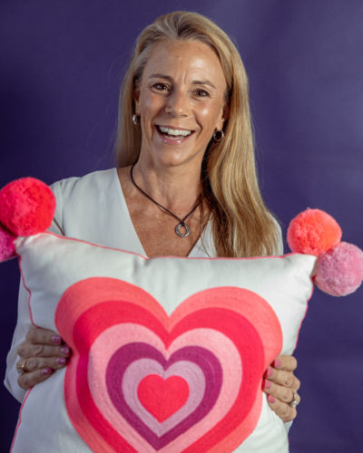 A blond woman in a white shirt holding up a pillow with a large pink heart.