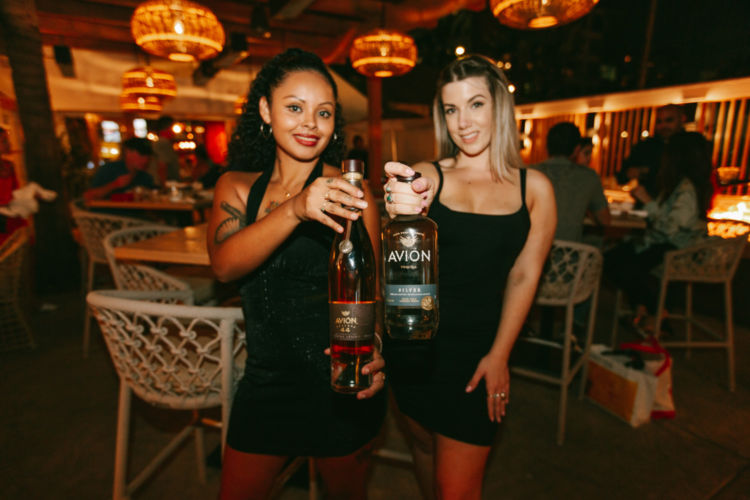 Two women wearing black dresses holding up two bottles of the Avion.