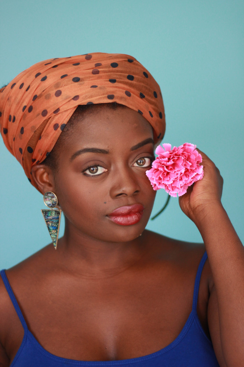 A woman in a blue shirt, statement earrings, orange hair turban posing with a pink flower.