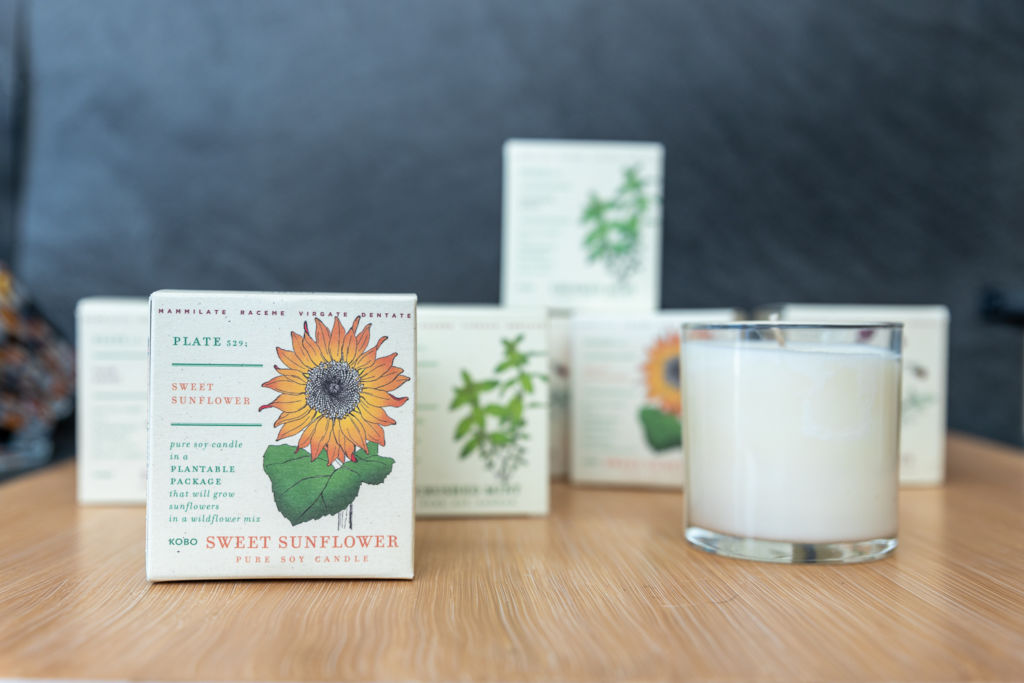 A white candle is photographed with its box with a sunflower printed on it.